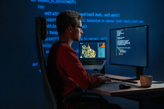 Data scientist writing code for artificial intelligence project, with programming scripts visible across desktop monitors. Coder in glasses working in front multiple screens at night. Copy space