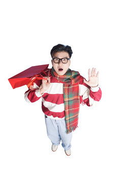 Shocked Shopper: A young man in festive holiday attire, complete with glasses and a scarf, reacts with wide-eyed shock and raised hands while clutching a bright red shopping bag.