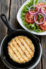 Fototapeta premium Top view of grilled provolone cheese in black cast iron pan. Fresh salad with sliced tomato, red onion, lettuce on white plate. Healthy vegetarian meal served on rustic wooden table for lunch dinner.