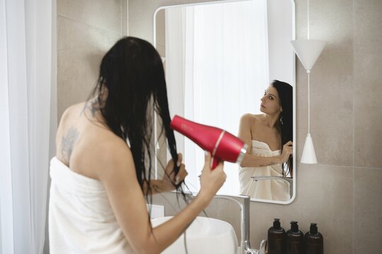 A woman stands in a bright, contemporary bathroom, holding a hair dryer and drying her wet hair. She is wrapped in a towel and gazes into the mirror, surrounded by minimalist decor