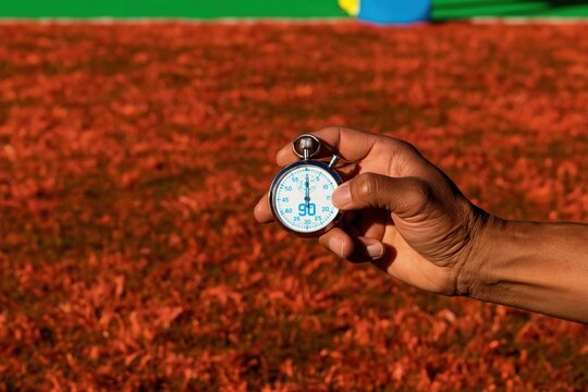 Hand of man holding stopwatch with minute display on a football field, featuring warm amber grass for sports and time management
