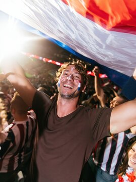 Paraguay football fan with painted face holding a rippled flag overhead in a crowded stadium, natural and euphoric for sports and national pride