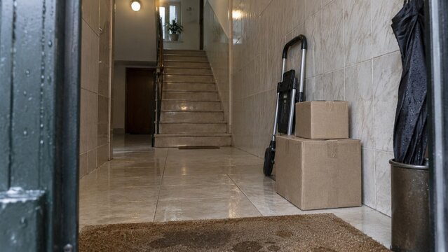 Two brown cardboard boxes sit on a tiled floor beside a metal hand truck, with a staircase and a closed door visible in the background hallway