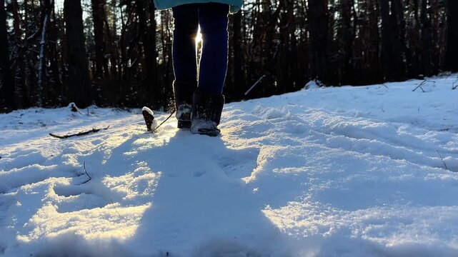 Legs of young girl in uggs hiking through snowy forest trail with sunlight at background. Female foot stepping on snow path in park at cold sunny day. Concept of winter active recreation.