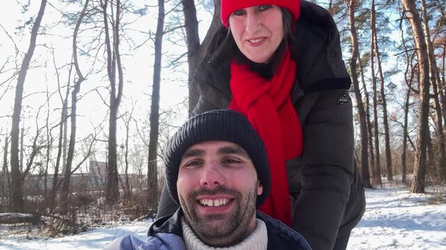 Happy woman push a sled with her man on snowy path in the sunny forest. Cheerful couple smiling at camera while pulling sledge at frosty woodland. Active pair enjoy fun winter walk outdoor. POV