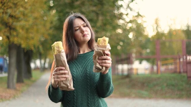 Hungry woman chooses small shawarma over large wrap in evening park. Green sweater and calorie conscious choice suggest mindful eating and dinner planning.