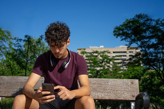 A young man sitting on a bench in a public park on a sunny day, scrolling through his smartphone with headphones around his neck.
