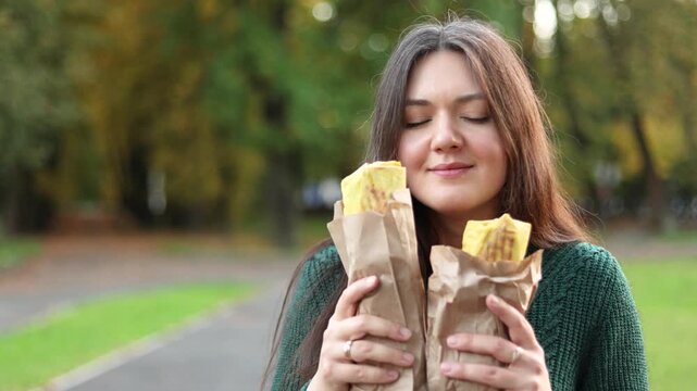 Young woman smells fresh shawarma in an autumn park and enjoys its aroma. Passing cars with headlights create a lively evening city background.