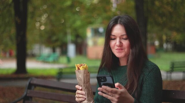Woman eats shawarma while browsing social media on smartphone in park. She combines a quick outdoor snack with digital activity during her busy day.