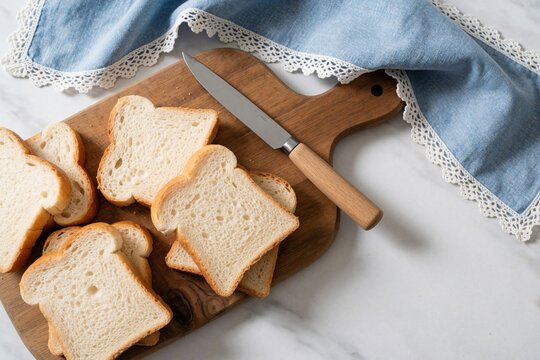 Top view of sliced white toast bread on wooden cutting board. Metal knife, blue linen napkin with lace edge lie on marble table. Fresh wheat slices ready for breakfast meal tasty sandwich snack in