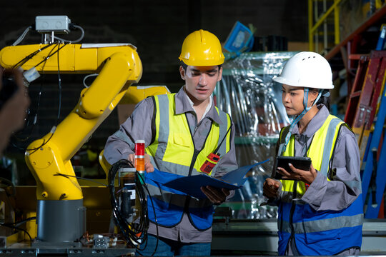 Multiethnic engineers examining an automated robotic arm with a tablet and clipboard discussing quality control and production efficiency in a high-tech manufacturing workshop.