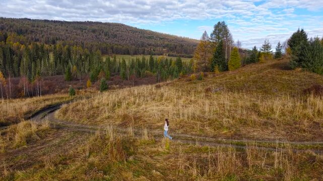 Drone high angle tracking shot from right to left following a woman walking on a rural mountain pass road in autumn.