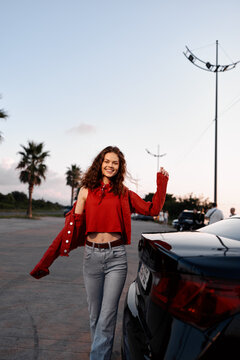 Young woman in a red cropped sweater and jeans stands beside a dark car in an outdoor parking lot, smiling under a clear evening sky with a soft glow and relaxed vibe.