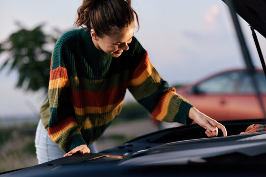 Young woman in colorful sweater checking car engine under open hood. Concept of breakdown, roadside assistance, and vehicle maintenance. Warm evening light.