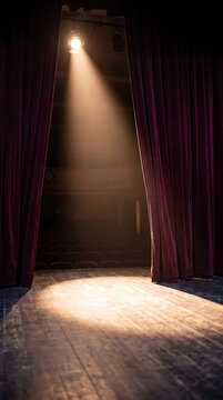 Empty theater stage framed by dark burgundy curtains with a warm spotlight shining across a wooden floor and rows of seats in the auditorium