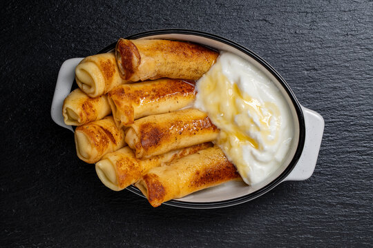 Crepes with sour cream and honey in white ceramic dish. Top view of thin rolled pancakes served with thick sour cream, yogurt and sweet honey syrup. Breakfast or dessert on black stone background