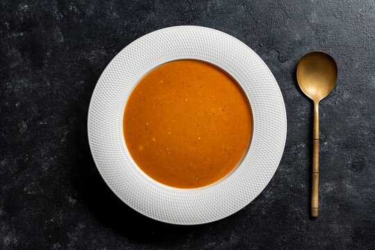 Pumpkin soup flat lay on dark background with golden spoon. Top view of orange cream soup in textured white plate. Minimalist healthy pumpkin or tomato puree food concept on black stone table