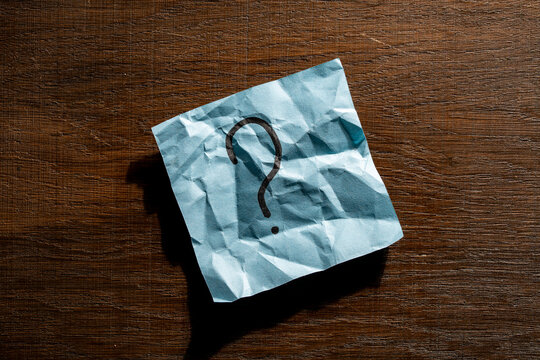 Question mark on crumpled blue paper note on wooden table. Crumpled blue sticky note with handwritten black question mark. Concept of confusion, doubts, choosing options and searching for answers
