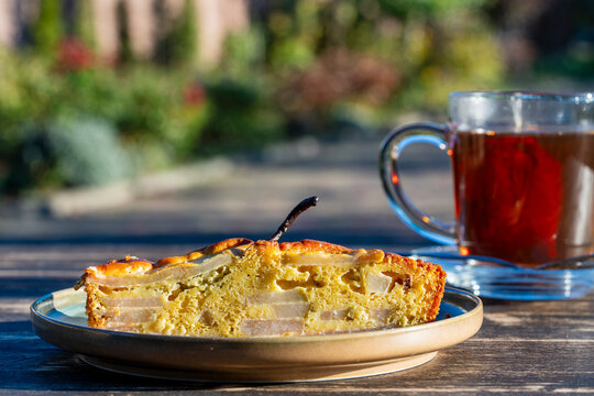 Pear pie slice on plate with tea cup in sunny garden. Closeup of homemade pear cake piece on wooden table. Sweet autumn fruit dessert served with black tea outdoors in sunny day