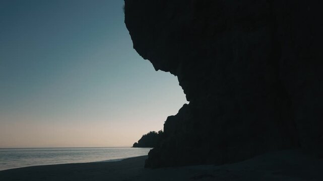 A quiet beach beneath a dimly gradient sky, with a massive rock formation jutting out in silhouette.