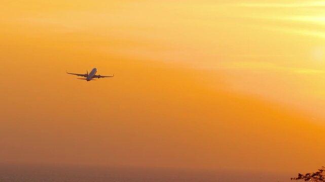 Passenger jet climbing after takeoff into orange sunset sky above sea, rear view silhouette, telephoto shot