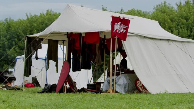 Red flag flutters above authentic medieval camping site in summer. Various colorful tunics and thick armor dry outdoors near warm cooking fire and large shield.
