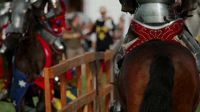 Close up shows armored knight on brown horse from behind. Blurry background reveals another armed rider preparing for jousting match behind wooden fence at tournament.
