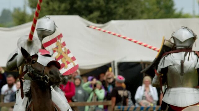Long wooden lance misses rival shield during rainy jousting clash. Blurred crowd watches medieval knights fighting on horseback near white festival tents in bad weather.