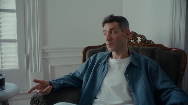 Male patient sitting in vintage armchair, looking aside and speaking during counseling session in psychology office