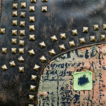 Close-up of distressed dark brown leather surface decorated with rows of metallic pyramid studs, adjacent to a section of frayed fabric featuring printed text and a small green patch