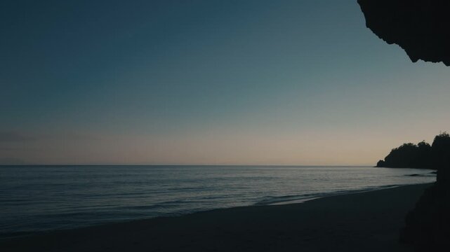 A quiet beach beneath a dimly gradient sky, with a massive rock formation jutting out in silhouette.