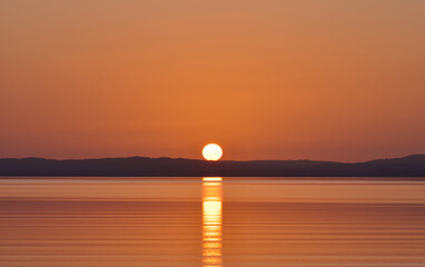 Beautiful golden sunset over calm coastal waters, with the setting sun casting a bright reflection across the sea and a dark distant shoreline beneath a warm orange sky. © romansucho 67