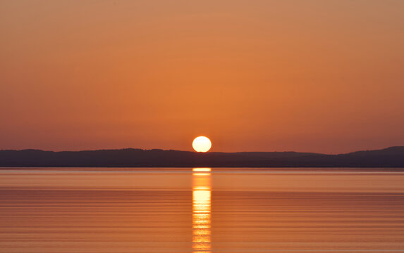 Beautiful golden sunset over calm coastal waters, with the setting sun casting a bright reflection across the sea and a dark distant shoreline beneath a warm orange sky.