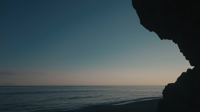 A quiet beach beneath a dimly gradient sky, with a massive rock formation jutting out in silhouette.