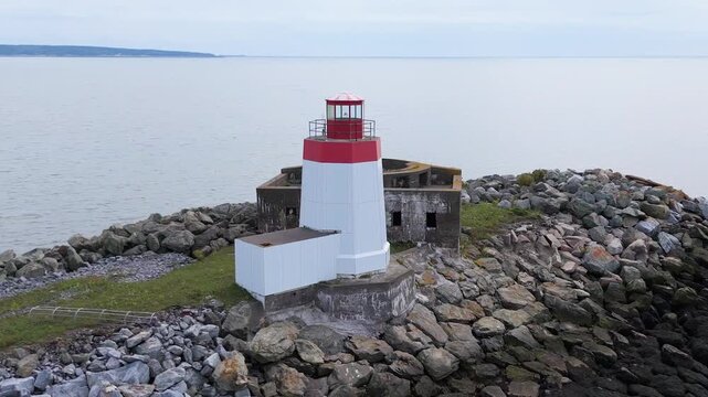 Areal view climbing away from an old lighthouse and the ruins of a gun emplacement. They rest on a narrow causeway built from boulders. Overcast day.