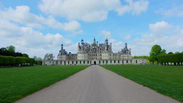 Renaissance castle at the end of a long straight avenue between green lawns and clipped tree rows under big cumulus clouds in spring, zoom in, Chateau de Chambord, Loire Valley, France