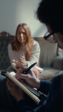 Professional psychotherapist writing notes in notebook with pen as listening to female patient explaining mental health issues during consultation. Vertical close-up shot