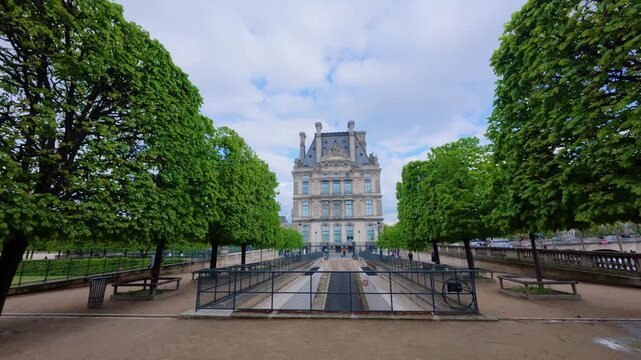 POV walk across the Tuileries Garden past rain puddles reflecting the trees toward the Pavillon de Flore of the Louvre between rows of trimmed trees on a cloudy spring day, Paris, France
