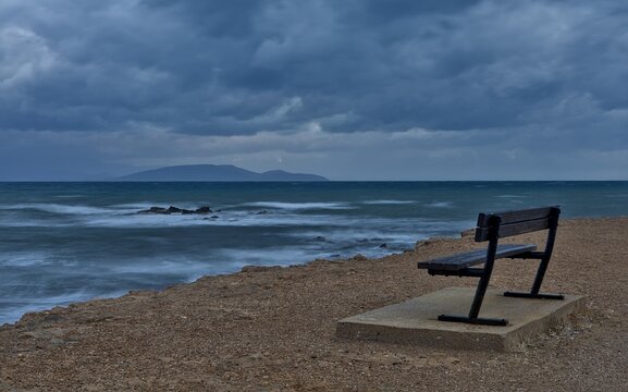 Empty seaside bench overlooking rough sea and Evia under dramatic clouds, Artemida, Greece