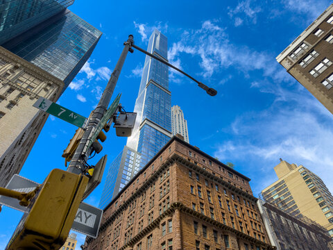 A streetlight crosses the view of a skyscraper above a Manhattan corner. A yellow signal sits below green signage as clouds pass between glass and brick facades.
