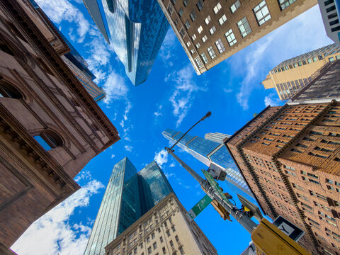 A ring of Manhattan buildings encloses the sky above a streetlight. The upward view places glass towers and masonry corners around clouds and green signage.
