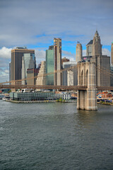 Fototapeta premium A stone tower of Brooklyn Bridge stands beside the Manhattan skyline. Suspension cables cross glass facades above a pier building and gray river water under clouds.