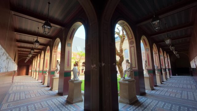 Slow walk through a painted cloister arcade with hanging lanterns and white statues on pedestals revealing a sunlit courtyard with a pruned tree and a fountain, Ecole des Beaux Arts, Paris, France