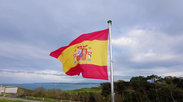 Spanish national flag with coat of arms waving in slow motion on a flagpole above a green headland at Cabo Mayor with the sea and coast beyond under a grey cloudy sky, Santander, Cantabria, Spain