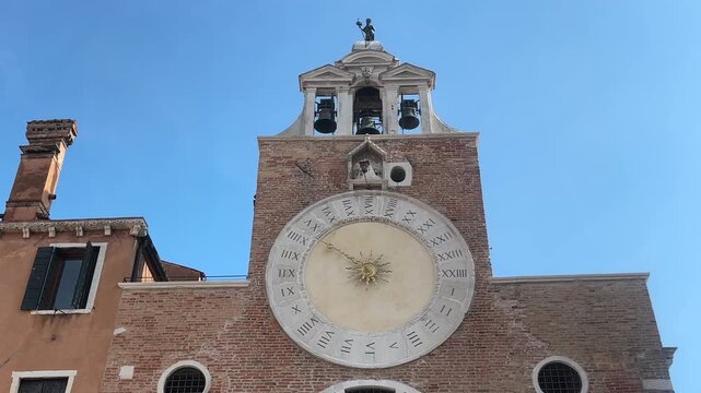 Low angle video zooming in on the historic large 24-hour clock face and bell tower of San Giacomo di Rialto church against a clear blue sky in Venice, Italy.