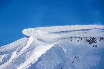 A massive overhanging snow cornice crowns a mountain summit as wind blows spindrift across its edge under a blue sky © Kafi
