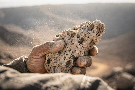 Close-up of a worker's dusty hand holding a porous volcanic rock in an arid desert landscape