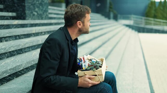 Dismissed desk worker sitting alone on stairs beside office box processing layoff. Career disruption shock