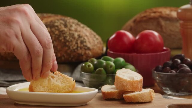 Bread dipping in olive oil close up, hand dips rustic bread into extra virgin olive oil on Mediterranean table with tomatoes and olives, Italian and Spanish food