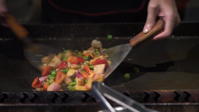 A dynamic close-up captures the vibrant colors of sliced peppers, onions, and garlic being tossed in a large wok with metal spatulas. The scene conveys heat and movement, highlighting the textures of